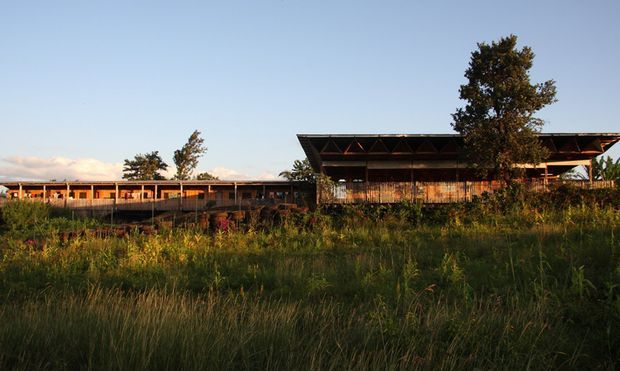 A low-rise, elongated building complex set within tall grasses and natural landscaping. The structure features dark timber or metal framing, extensive horizontal glazing, and a dramatic broad overhanging roof with exposed triangular structural elements. The image is an exterior architectural view photographed in warm late-day light, with a rural or landscaped setting.