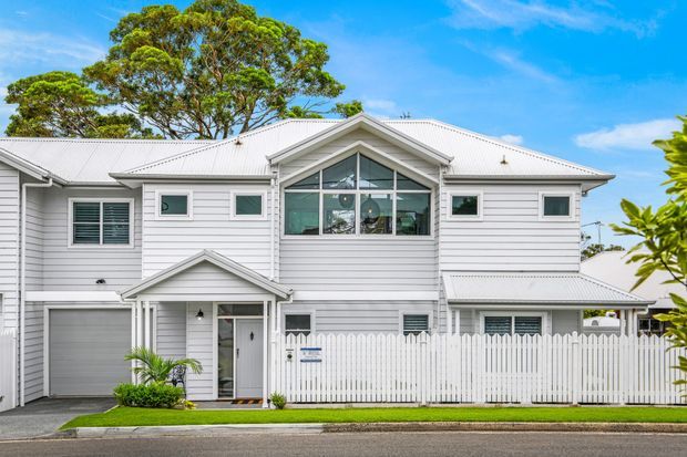 Front exterior view of a large two-storey white weatherboard home with a light grey corrugated metal roof. The facade features multiple gables, projecting eaves, white-framed windows, a central upper-level glazed feature, an attached garage, covered entry porch, and a white picket fence. Mature trees and a suburban street setting surround the property.