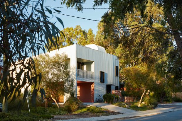 A contemporary two-storey residence viewed from the street, set among mature eucalyptus and landscaped planting. The compact facade combines pale vertically ribbed cladding, deep recessed balcony openings, dark-framed windows, a warm timber-toned garage door, and sections of exposed red brick. A concrete path connects the home to the street, creating a suburban Australian streetscape.