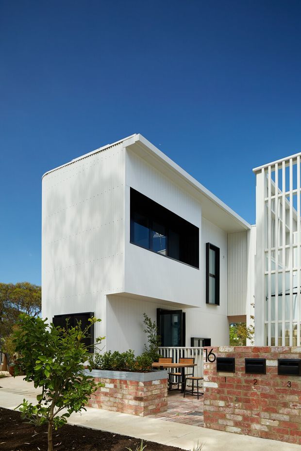 Exterior architectural photograph of a crisp white contemporary multi-storey residence with a flat roof, projecting upper volume, black-framed windows, and a tall white vertical-screen facade element. The house is set behind low red brick retaining and mailbox walls, with a paved entry courtyard, planted garden beds, and a clear blue sky.