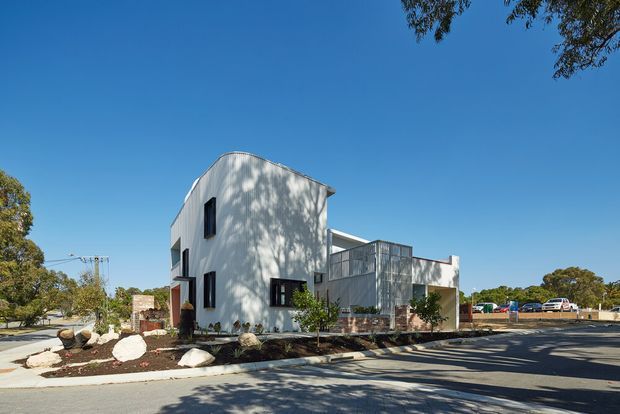 A contemporary multi-level residential building viewed from the street on a sunny day. The facade combines smooth white rendered walls with dark-framed windows and a projecting upper form, while a glazed or screened central section and pale masonry base add visual contrast. A landscaped front setback with rocks and sparse planting separates the home from the paved street and driveway.