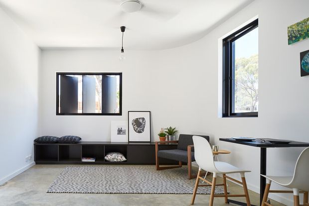 Bright, compact contemporary interior combining a living area and dining/workspace. The room has white walls and ceiling, polished concrete flooring, a low dark built-in storage unit, a patterned area rug, a dark lounge chair, white dining chairs, and a slim dark table. Black-framed windows provide abundant natural light, with simple artwork and potted plants adding restrained decoration.