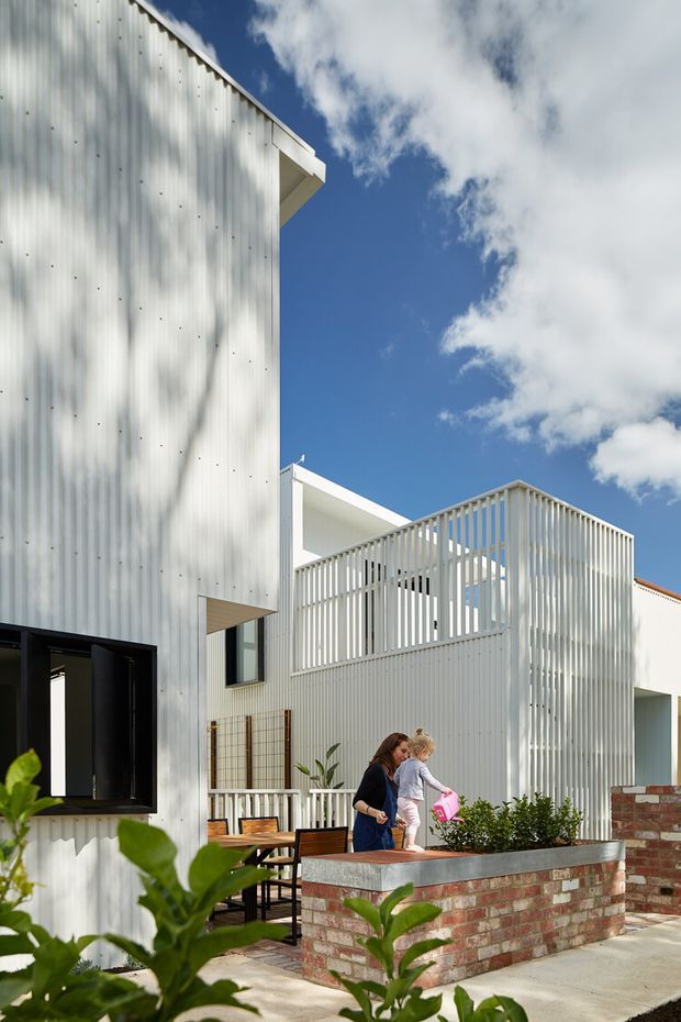 Exterior view of a contemporary multi-level residence with a bright white corrugated metal-clad facade, projecting roof forms, and a screened upper terrace enclosed by vertical white slats. A ground-level courtyard or patio includes outdoor dining furniture, raised brick planters with concrete caps, and landscaping. The image is photographed in strong daylight against a blue sky, with people present in the courtyard.
