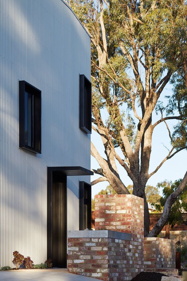Exterior view of a contemporary two-storey residence with a tall white vertically ribbed metal-clad facade, narrow black-framed windows, and a projecting dark entrance canopy. Curved and stepped walls of exposed variegated brick define the front boundary, set among mature eucalyptus trees and landscaped paving. The composition emphasises the contrast between crisp modern cladding and warm brickwork in a suburban natural setting.