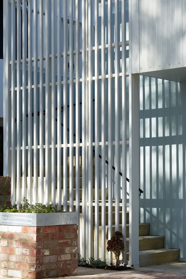 Exterior view of a contemporary multi-level residential entrance, screened by closely spaced vertical metal fins. The fins create strong linear shadows across a pale rendered wall and stairwell, with exposed brick planter walls and low landscaping in the foreground. A partially visible internal staircase and entry steps sit behind the open screen.