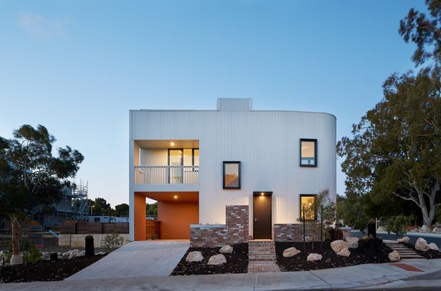 Exterior view of a contemporary multi-level residence at dusk, featuring a curved corrugated metal-clad facade, recessed balcony and carport-like ground-level opening, dark-framed windows, a timber front door, and a brick feature wall. The house is set behind a landscaped frontage with dark mulch, large rocks, paving, and mature trees.