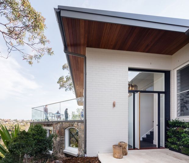 Exterior view of a contemporary residential entry, featuring white painted brick walls, a dark metal-edged roof with a warm timber-lined soffit, and a tall glazed doorway. A stone retaining wall and glass-balustraded terrace are visible beside the entry, with landscaped planting in the foreground.