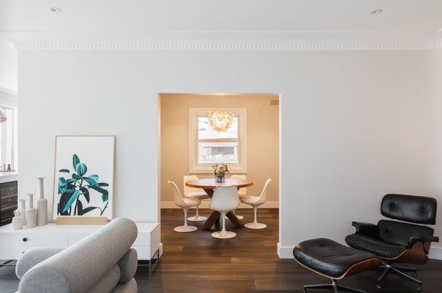 Bright, refined open-plan living area with a contemporary grey sofa in the foreground, a black leather lounge chair and ottoman, white built-in cabinetry, and a framed botanical artwork. Through a wide opening, a compact dining area features a round white table, sculptural white chairs, timber flooring, and a warm-toned wall with a window and chandelier. Decorative ceiling cornice adds a traditional architectural detail.