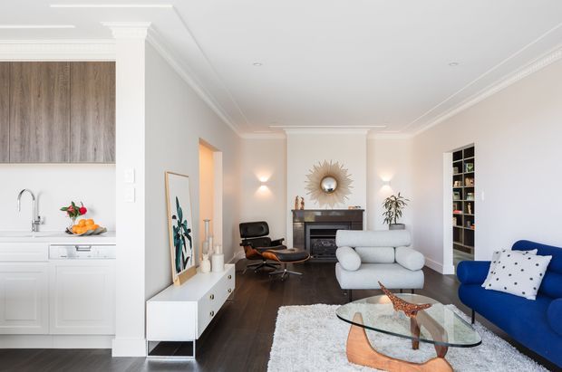 Bright open-plan living room with a white kitchen at left and a refined sitting area centred on a dark fireplace and sunburst mirror. The room features dark timber flooring, white walls and cabinetry, a sculptural glass-and-timber coffee table, a white sofa, blue armchairs, built-in shelving, wall sconces and decorative objects. Traditional ceiling cornices and trim contrast with contemporary furnishings.