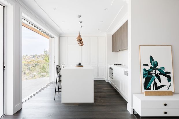 Bright contemporary white kitchen with a central island, white Shaker-style panelled cabinetry, dark timber flooring, and a warm wood overhead cabinet. Full-height sliding glazing opens to an exterior view, while a framed botanical artwork and white storage console sit in the foreground. The room has a clean, refined look with subtle natural timber accents and pendant lighting.