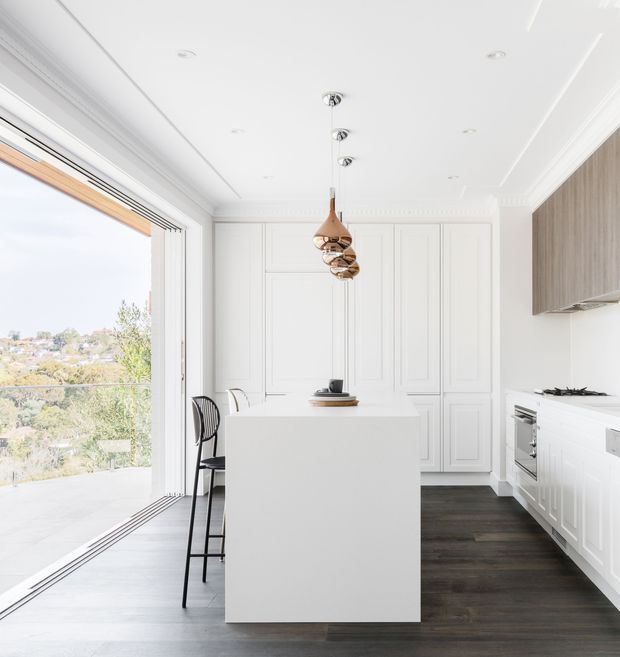Bright, refined kitchen interior with a large white island, extensive white raised-panel cabinetry, dark timber flooring, and a bank of integrated appliances. A trio of sculptural pendant lights hangs above the island, while full-height glazed doors open to a balcony or terrace with a leafy suburban view. Decorative ceiling cornices and wall panelling add a classic architectural character.