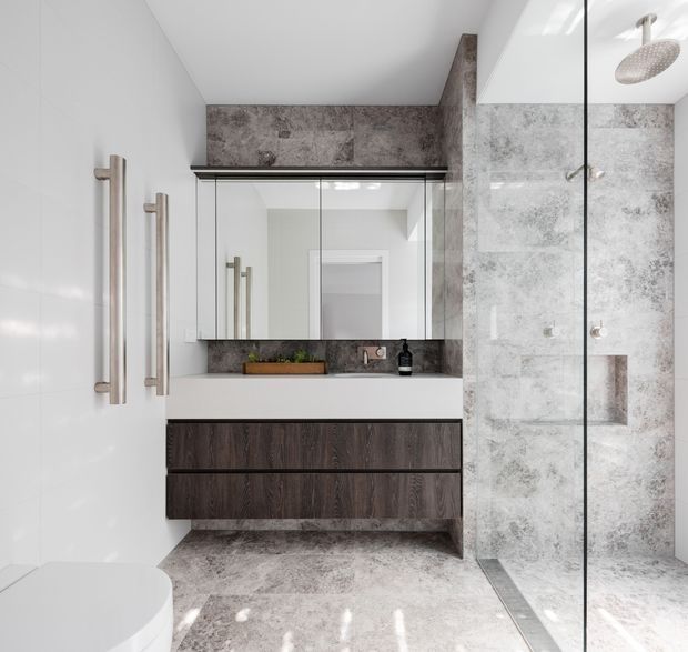 Contemporary bathroom with a floating dark timber vanity, white integrated basin, mirrored wall cabinetry, and large-format mottled grey stone-look wall and floor surfaces. A frameless glass shower enclosure with a round overhead shower occupies the right side, while a white toilet is partially visible at the left. Brushed metal vertical handles and fittings add a restrained modern detail.