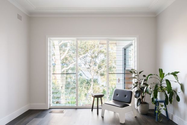 Bright, serene sitting area with a white moulded ceiling and cornice, pale neutral walls, dark timber flooring, and a large white-framed glazed opening with horizontal louvres or blinds. A sculptural white lounge chair and small stool sit beside several leafy indoor plants, creating a clean, airy interior with a view to greenery outside.