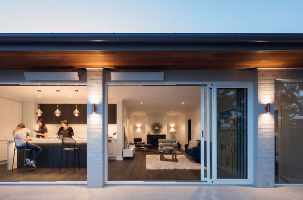 Dusk exterior view of a contemporary single-storey home with a covered timber-lined patio or verandah. Large glazed openings reveal an open-plan kitchen with a breakfast island on the left and a living room with sofas and a rug in the centre. White-painted brick columns and walls, dark roof trim, timber soffit lining, recessed ceiling lights, and wide sliding glass doors define the facade.