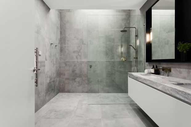 Contemporary bathroom with large-format grey stone-look wall and floor tiles, a spacious walk-in shower with frameless glass panel and exposed shower fittings, a heated towel rail, and a floating white vanity with a grey stone benchtop. A large black-framed mirror and dark cabinetry add contrast.
