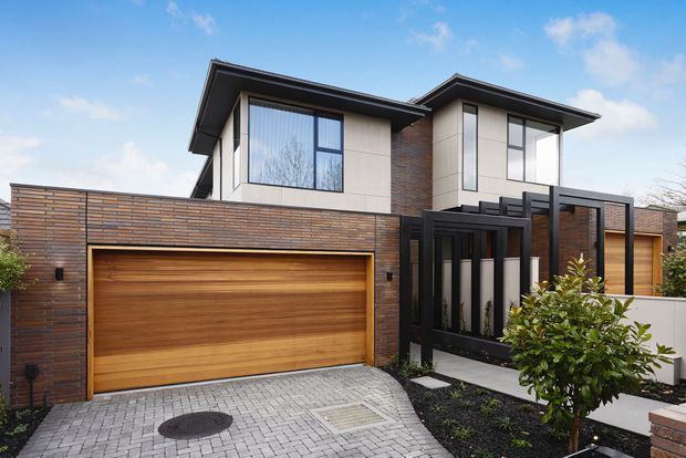 Contemporary two-storey residential facade with a projecting dark roofline, large vertical windows, pale rendered upper walls, exposed brick detailing, and a timber garage door integrated into a brick boundary wall. A black-framed entry screen and landscaped front garden sit beside a paved driveway.