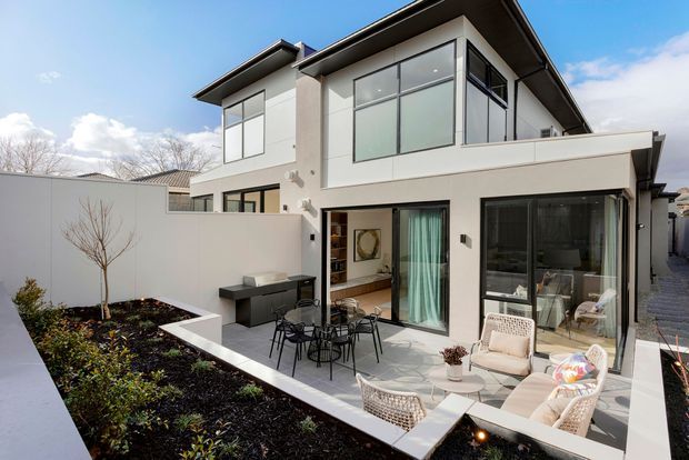 Bright exterior view of a contemporary two-storey residence with a flat roof, crisp white rendered walls, extensive dark-framed glazing, and a raised upper-level volume. A paved courtyard patio with outdoor dining and lounge furniture sits beside landscaped garden beds, while the interiors are partially visible through the large glass doors.