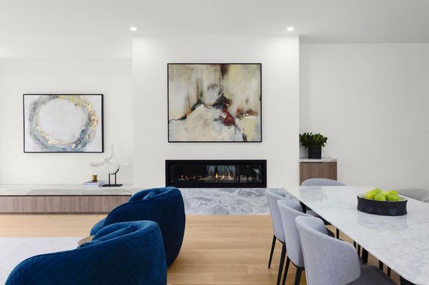 Bright, contemporary open-plan living and dining area with pale timber flooring, a long white marble dining table, upholstered dining chairs, deep blue lounge chairs, and a linear built-in fireplace set into a white feature wall. Abstract artwork, a low timber media console, recessed ceiling lighting, and restrained decor create a refined modern interior.