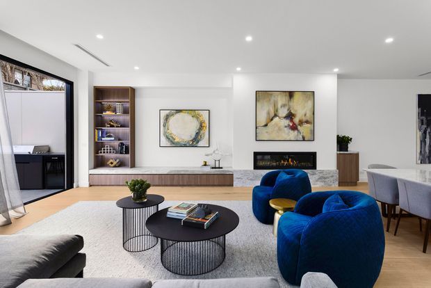 Bright contemporary living room with white walls and recessed ceiling lights, a large pale rug, black round coffee tables, a grey sectional sofa, and two vivid blue curved lounge chairs. Built-in timber shelving and low cabinetry line the rear wall beside abstract artwork and a linear fireplace. A dining area and desk are visible at the edges, with large dark-framed glazing on the left.
