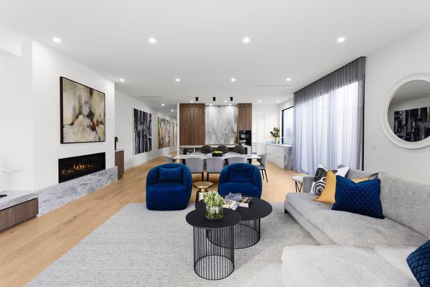 Bright contemporary open-plan living and dining room with light timber flooring, a large pale sectional sofa, deep blue lounge chairs and cushions, black round coffee tables, a marble-look fireplace surround, large artwork, and floor-to-ceiling sheer curtains. A dining area and kitchen are visible beyond the living space.