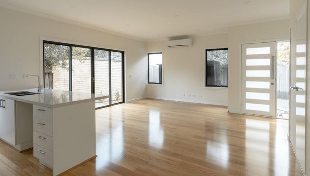Bright open-plan interior combining a compact kitchen with a spacious living or dining area. The room has polished light timber flooring, white walls and cabinetry, large glazed sliding doors opening to a courtyard, and a contemporary white entry door with horizontal glass panels. The space is largely unfurnished and receives abundant natural light.