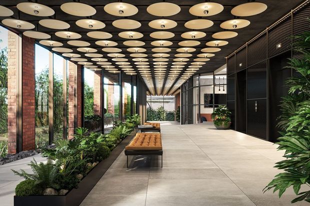 A sophisticated, elongated commercial interior hallway or lobby with a dark ceiling covered in repeated oval pendant lights, tall glazed openings between exposed brick columns, integrated indoor planting beds, pale large-format floor tiles, and dark metal mesh joinery along one side. A central upholstered bench provides seating, while lush greenery and daylight create a refined indoor-outdoor atmosphere.