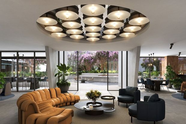 A spacious, refined contemporary living room or lounge with floor-to-ceiling glazing, sculptural angled columns, and an expansive circular ceiling feature containing multiple pendant lights. Curved caramel sofa seating and dark rounded lounge chairs surround layered circular coffee tables. The room opens toward a planted outdoor terrace and includes a dining or hospitality area to the side, creating an airy indoor-outdoor connection.