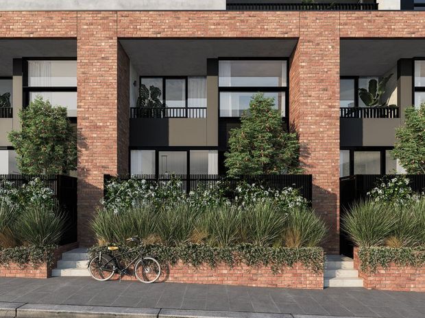 Street-facing view of a contemporary multi-level townhouse or apartment facade. The building features warm red brick masonry, deep recessed entrances and balconies, black metal balcony railings, dark-framed glazing, and landscaped planter beds with ornamental grasses and shrubs along the pavement.
