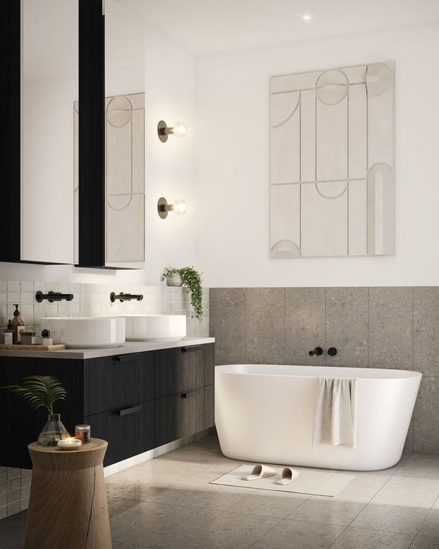 A refined contemporary bathroom featuring a double vanity with dark vertical-groove cabinetry, white vessel basins, wall-mounted black tapware, a freestanding white bathtub, and large grey stone-look wall and floor tiles. A large mirror, warm globe sconces, geometric wall artwork, and restrained decorative styling create a calm, modern atmosphere.