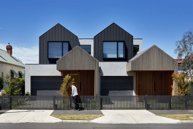 Street-facing view of a contemporary multi-storey residential duplex or townhouse development. The facade combines two dark, gabled upper volumes with large projecting windows, pale rendered walls, black garage doors, and strongly textured vertical timber-look screening around the entry and garage fronts. A dark vertical-slat fence and concrete footpaths occupy the foreground.