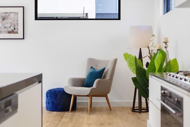 Bright, refined living area with a mid-century-inspired upholstered armchair, blue ottoman and cushion, floor lamp, indoor plant, framed artwork, and pale timber flooring. A built-in kitchen or cabinetry edge is visible in the foreground, while high-set windows bring in natural light.