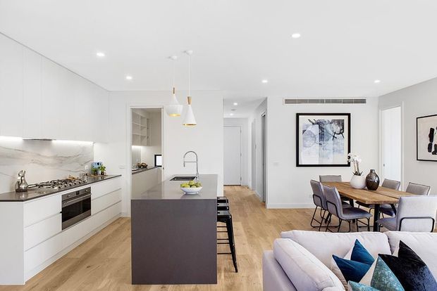 Bright open-plan contemporary kitchen, dining and living area with white flat-panel cabinetry, a dark central island, pale timber flooring, marble-look splashback, pendant lights and neutral furnishings. The kitchen occupies the foreground, with the dining and living areas extending behind it.