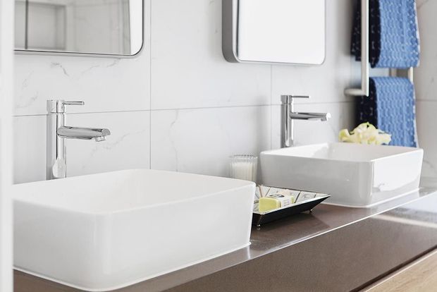 Bright contemporary bathroom vanity with two white rectangular vessel basins, wall-mounted mirrors, chrome single-lever faucets, large white marble-look wall tiles, and a dark stone-look countertop. Blue towels and small bathroom accessories provide subtle colour accents.