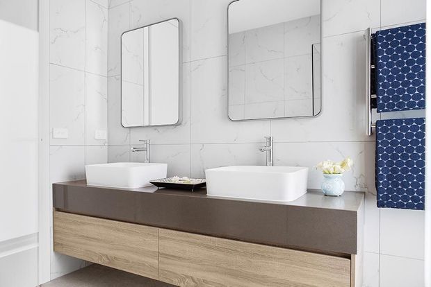 Contemporary double-vanity bathroom with two white vessel basins on a dark benchtop, light timber floating cabinetry, large rounded-corner mirrors, and pale marble-look wall tiles. A blue patterned towel adds a strong accent.