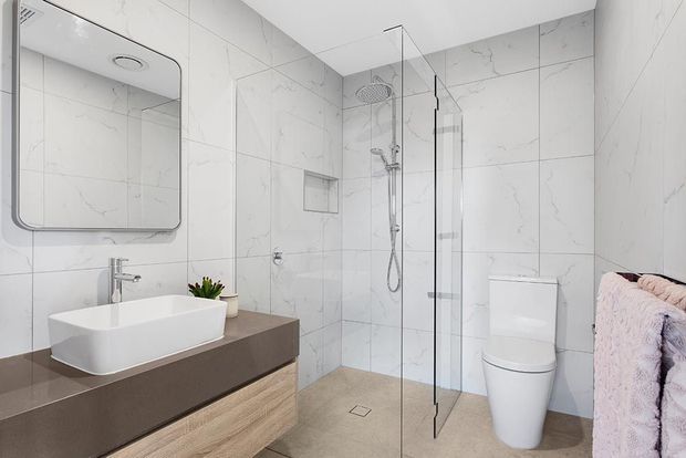 Contemporary bathroom with large white marble-look wall tiles, a frameless glass walk-in shower, wall-mounted rain shower and handheld fittings, a white vessel basin on a dark stone-look benchtop, and light timber vanity cabinetry. A toilet, mirrored cabinet, recessed shower niche, and soft pink towel are also visible.
