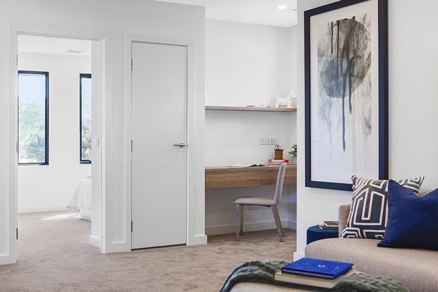 Bright, contemporary bedroom or living area with soft grey carpet, white walls and doors, a built-in timber desk with shelving, a transparent chair, and a partially visible upholstered sofa. A large framed abstract artwork and blue-and-patterned cushions add contrast. An open doorway reveals a secondary bedroom with a bed and tall windows.
