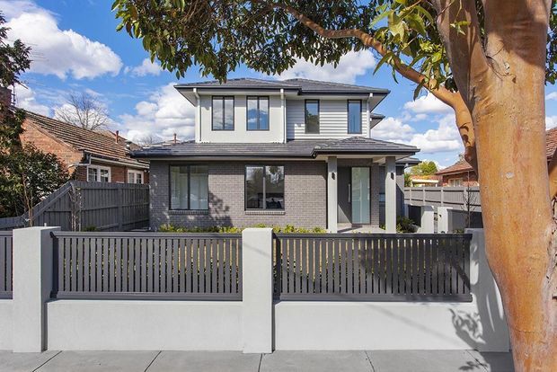 Front exterior view of a contemporary two-storey suburban residence, with a grey brick and horizontal weatherboard facade, low-pitched roof, dark-framed windows, and a private front fence and gate. Mature trees and neighbouring homes frame the street-facing elevation.