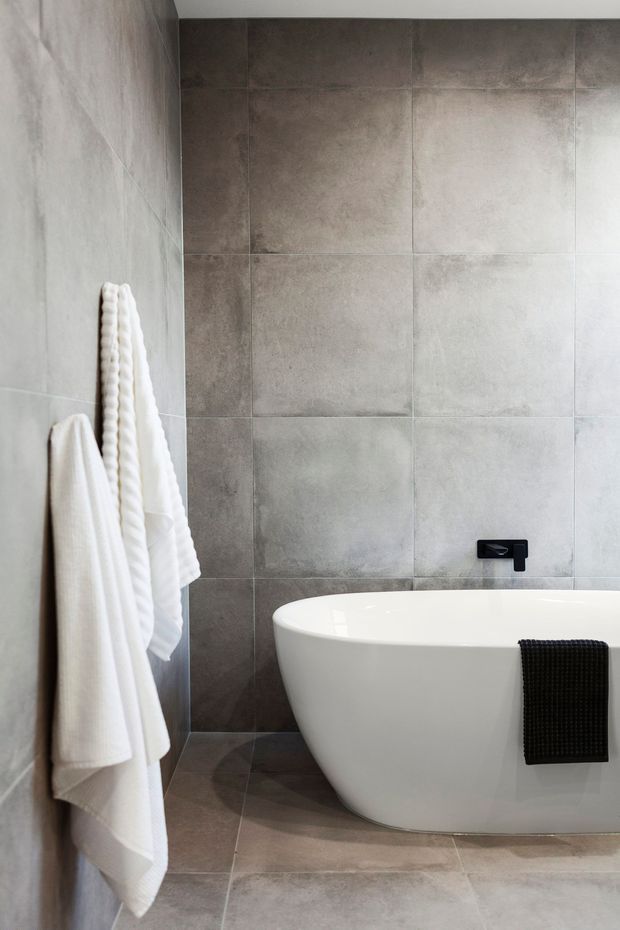 A contemporary bathroom featuring a freestanding white bathtub against large-format grey stone-look tiled walls and flooring. A black wall-mounted tap and black towel contrast with the neutral finishes, while white towels hang on the left wall.