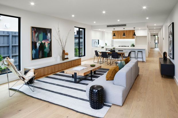 A bright, open-plan contemporary living room flowing into a dining area and kitchen. The space features pale timber flooring, white walls and cabinetry, a long light-grey sectional sofa, black-and-white striped rug, timber coffee table, sculptural lounge chair, black pendant lights, and a warm timber kitchen island backdrop. Large windows and recessed ceiling lights create an airy, refined interior.
