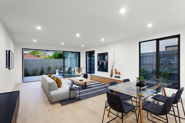 Bright, contemporary open-plan living and dining room with pale timber flooring, white walls, recessed ceiling lights, large black-framed windows and glazed sliding doors opening to a fenced outdoor area. A grey sofa, dark striped rug, timber coffee table and glass dining table with dark upholstered chairs create a clean, refined interior.