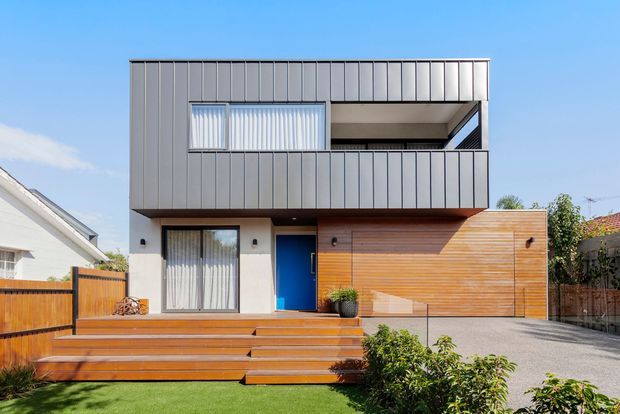 Front exterior view of a contemporary two-storey residence with a boxy flat-roof form, charcoal vertical metal cladding, white rendered lower walls, warm horizontal timber-clad garage frontage, and a broad timber deck with steps leading to the entry. A recessed upper balcony and large glazed openings are visible.
