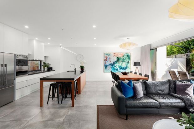 Bright open-plan kitchen, dining, and living area with a contemporary white kitchen, large island with timber breakfast bar, stainless-steel appliances, grey tiled flooring, dining table, and a black leather sectional sofa. Large sliding glass doors open to an outdoor area, while colourful artwork and pendant lighting add visual accents.