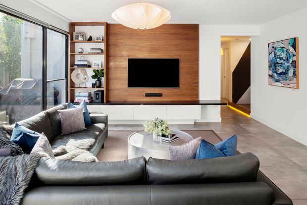 Contemporary living room with a large black leather sectional, neutral soft furnishings, a round coffee table, and a wall-mounted television set into a warm timber feature wall. Built-in white cabinetry and open shelving flank the entertainment unit, while full-height glazing brings in natural light. A sculptural pendant light, polished grey flooring, and colourful artwork complete the refined interior.