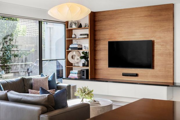 A contemporary living room featuring a large timber feature wall with integrated television, long white built-in cabinetry, open display shelving, a leather sofa, and a sculptural pendant light. Full-height glazing opens to a leafy outdoor area, creating a bright, refined interior.
