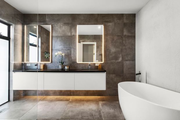 Contemporary bathroom featuring a long wall-mounted white vanity with dark stone benchtop, twin illuminated mirrors, large-format dark grey-brown tiled walls and floor, a freestanding white bathtub, and black-framed glazing. The space has a clean, refined, softly illuminated atmosphere.