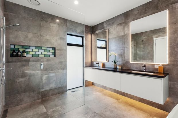 A contemporary bathroom with large grey concrete-look tiled walls and floor, a frameless glass walk-in shower, recessed shelf with patterned accent tiles, and a floating white double vanity with a dark benchtop. Large illuminated mirrors and warm under-vanity lighting add contrast to the minimalist fit-out.
