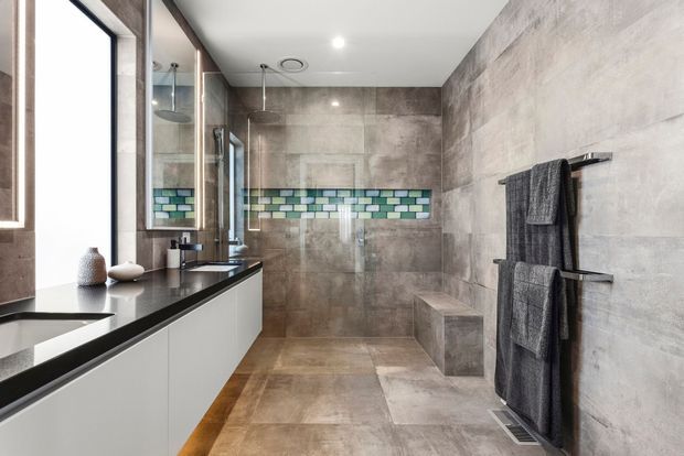 Contemporary bathroom with a long floating white vanity, dark stone-look benchtop, large grey-beige tiled walls and floor, frameless glass shower, recessed shower niche with green and blue mosaic tiles, and a wall-mounted heated towel rail. The room is bright, streamlined, and highly cohesive in neutral tones with a subtle coloured tile accent.