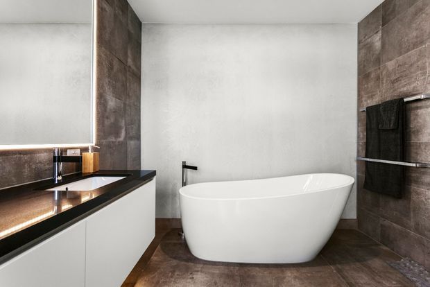 Contemporary bathroom featuring a freestanding white oval bathtub, dark stone-look wall and floor finishes, a floating white vanity with a dark benchtop, black tapware, and integrated warm mirror lighting. The space has a clean, refined, high-contrast aesthetic.