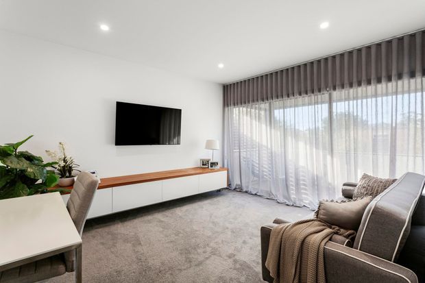 Bright contemporary living room with a neutral palette, low-profile white built-in media cabinetry, timber-toned console surface, wall-mounted television, large sheer-curtained glazing, grey carpet, and a comfortable upholstered sofa with layered cushions and throw.