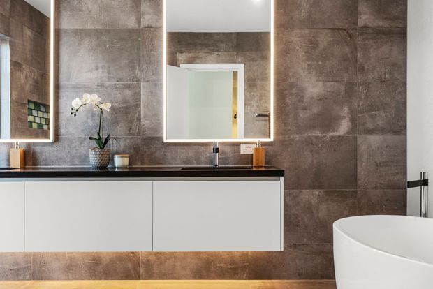Contemporary bathroom with large-format brown-grey tiled walls, a floating white vanity with black benchtop and integrated basin, illuminated mirrors, wall-mounted tapware, and a freestanding white bathtub. Warm timber flooring and restrained accessories add contrast.